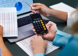 A person uses a calculator in front of a laptop in an office