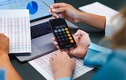 A person uses a calculator in front of a laptop in an office
