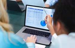 A woman points at a spreadsheet displayed on an office laptop