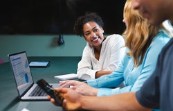 A smiling woman talks to her colleague in front of a laptop