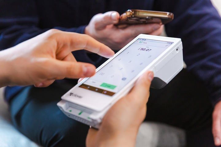 A close-up of someone holding the Clover Flex card machine with the right hand while touching the screen with the fingers of the left hand. In the background: a hand holding a phone.