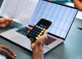 A person holds up a calculator in front of a laptop displaying a spreadsheet