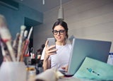 A young woman in a work studio is using her smartphone in front of a laptop