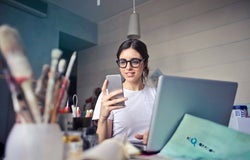 A young woman in a work studio is using her smartphone in front of a laptop