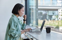 Millennial Asian student prepares for test or exam browses laptop studies in coffee house makes notes in notepad watches online webinar learns foreign language. E learning and online studying concept