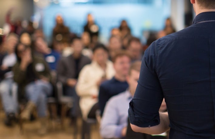 Back of a man presenting to a group of seated people in soft focus