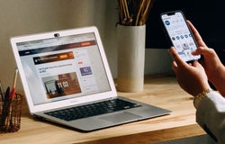 A person uses their smartphone to check something, while their laptop sits open on their desk