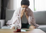 A man sitting on a grey sofa blowing his nose with a tissue