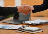 Two people shaking hands over a desk littered with notes and a calculator