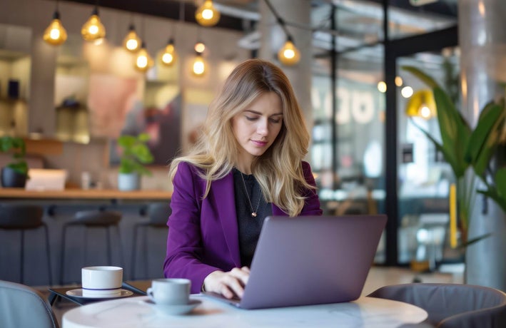A blonde woman with a purple blazer sitting at a cafe table working on a laptop.