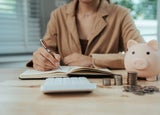 A person writes in a notebook, in front of a calculator and a piggy bank